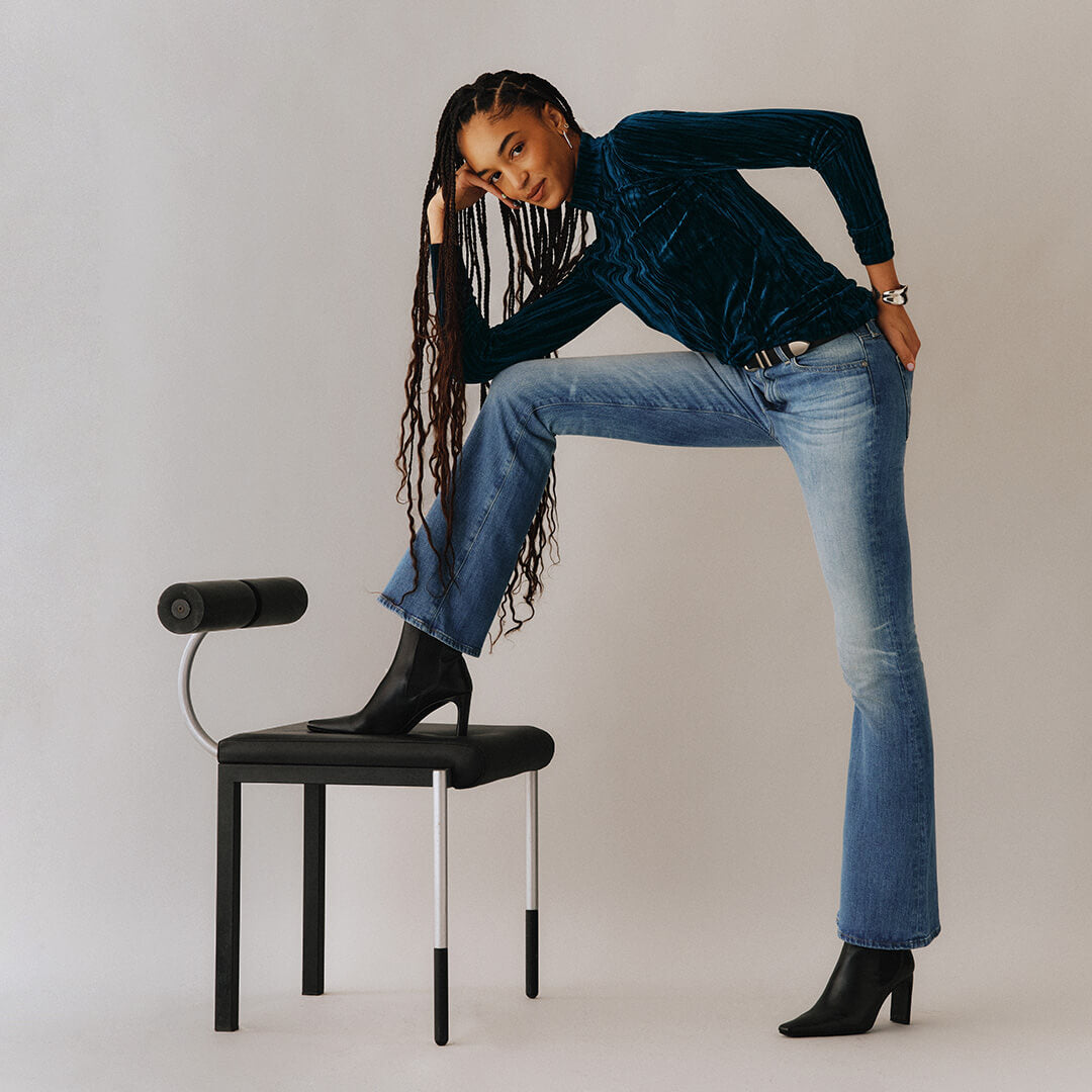Woman in blue velvet top and jeans posing on a chair against a plain background