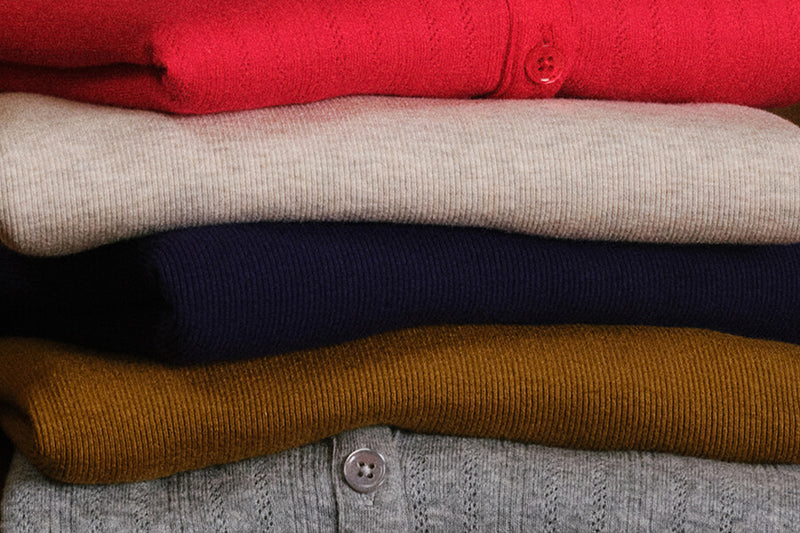 A close-up stack of folded knit cardigans in red, beige, navy, mustard yellow, and heather gray, each showing a button closure and ribbed texture.
