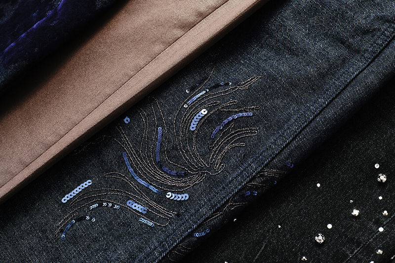 Close-up of a dark denim garment with embroidered blue and silver sequined wave details, layered over a brown suede-like fabric and a black fabric with rhinestone embellishments.