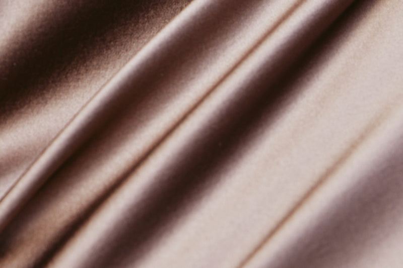 A close-up of draped satin-like fabric in a warm taupe-brown color, showing soft folds and a subtle sheen.