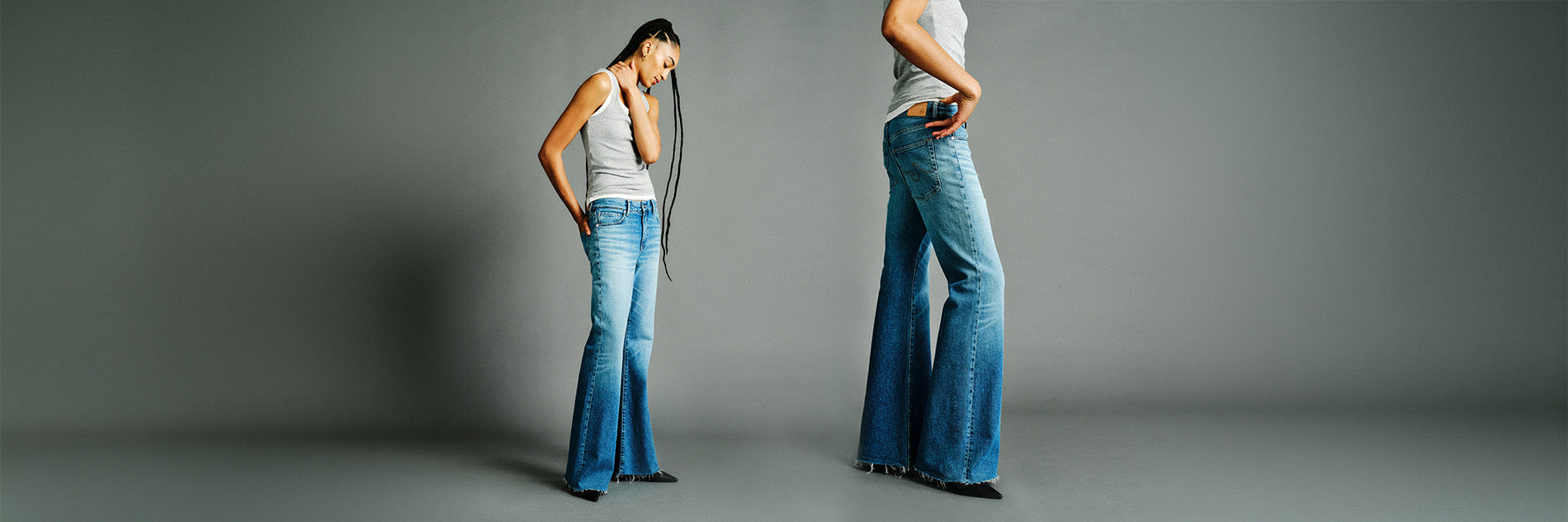 Model wearing blue flared jeans and white tank tops standing against a plain gray studio background