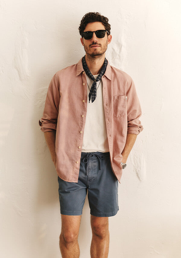 Man with sunglasses, pink shirt, and blue shorts on, standing in front of a white wall