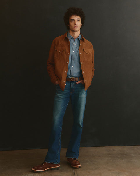 Man wearing a brown jacket, blue shirt, and jeans against a dark background