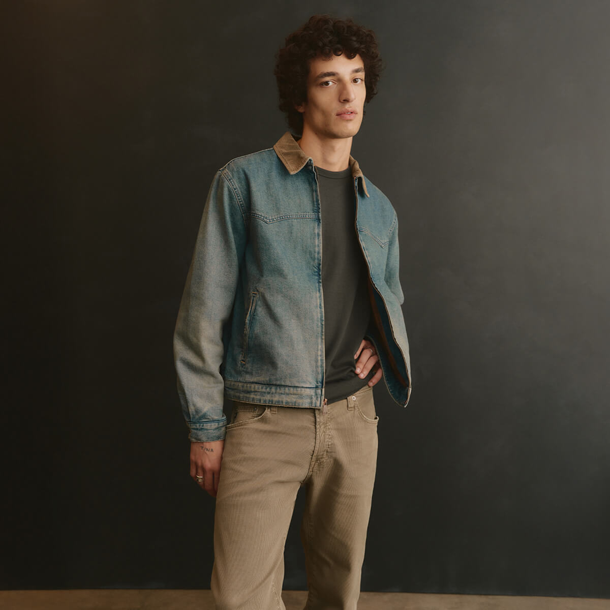 Man wearing a denim jacket and beige pants against a dark background