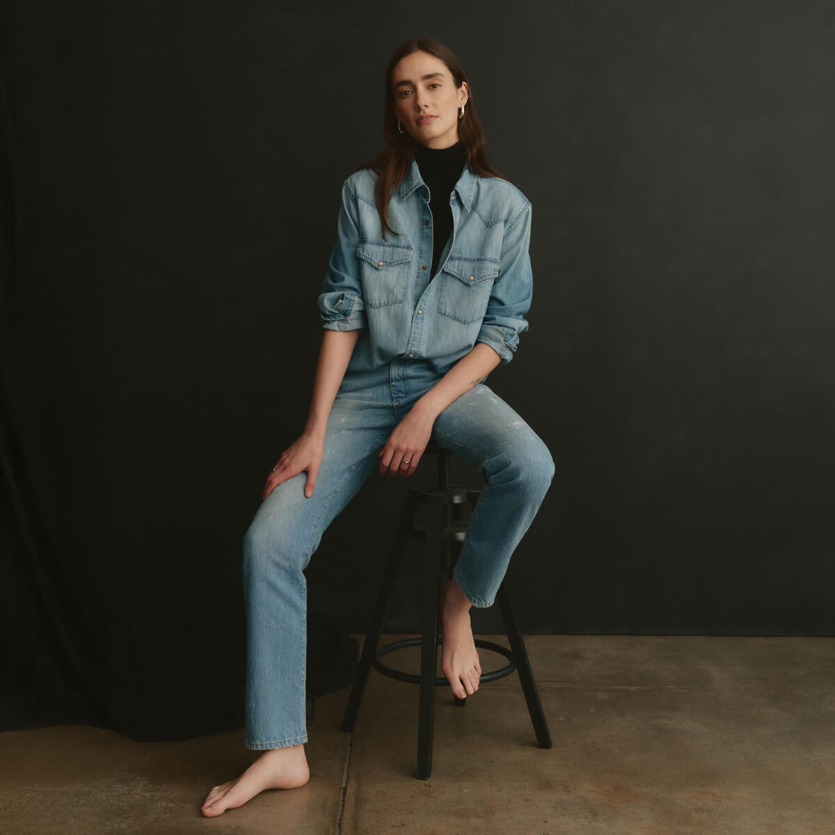 Woman wearing a denim outfit sitting on a stool against a black background