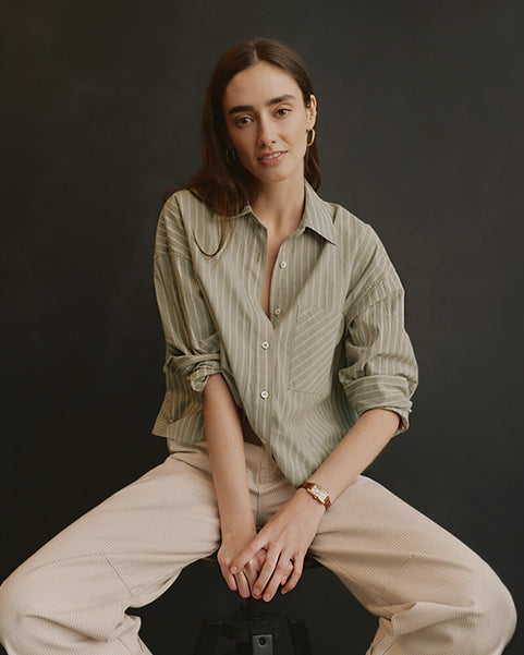 Woman wearing a light green striped shirt and beige pants sitting against a dark background