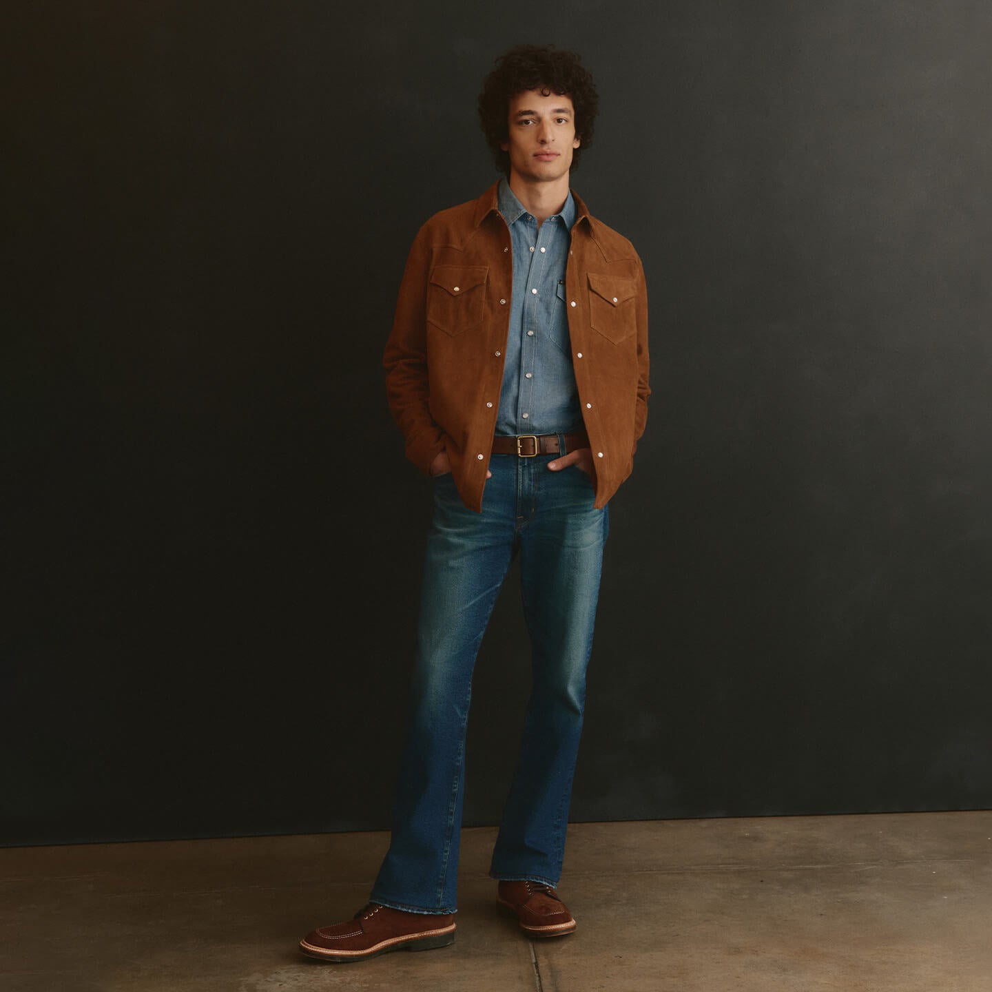 Man wearing a brown suede jacket, blue shirt, and jeans against a black background