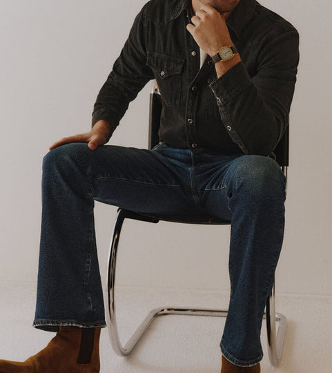 Man sitting on a chair wearing a dark denim jacket and jeans with a blurred background