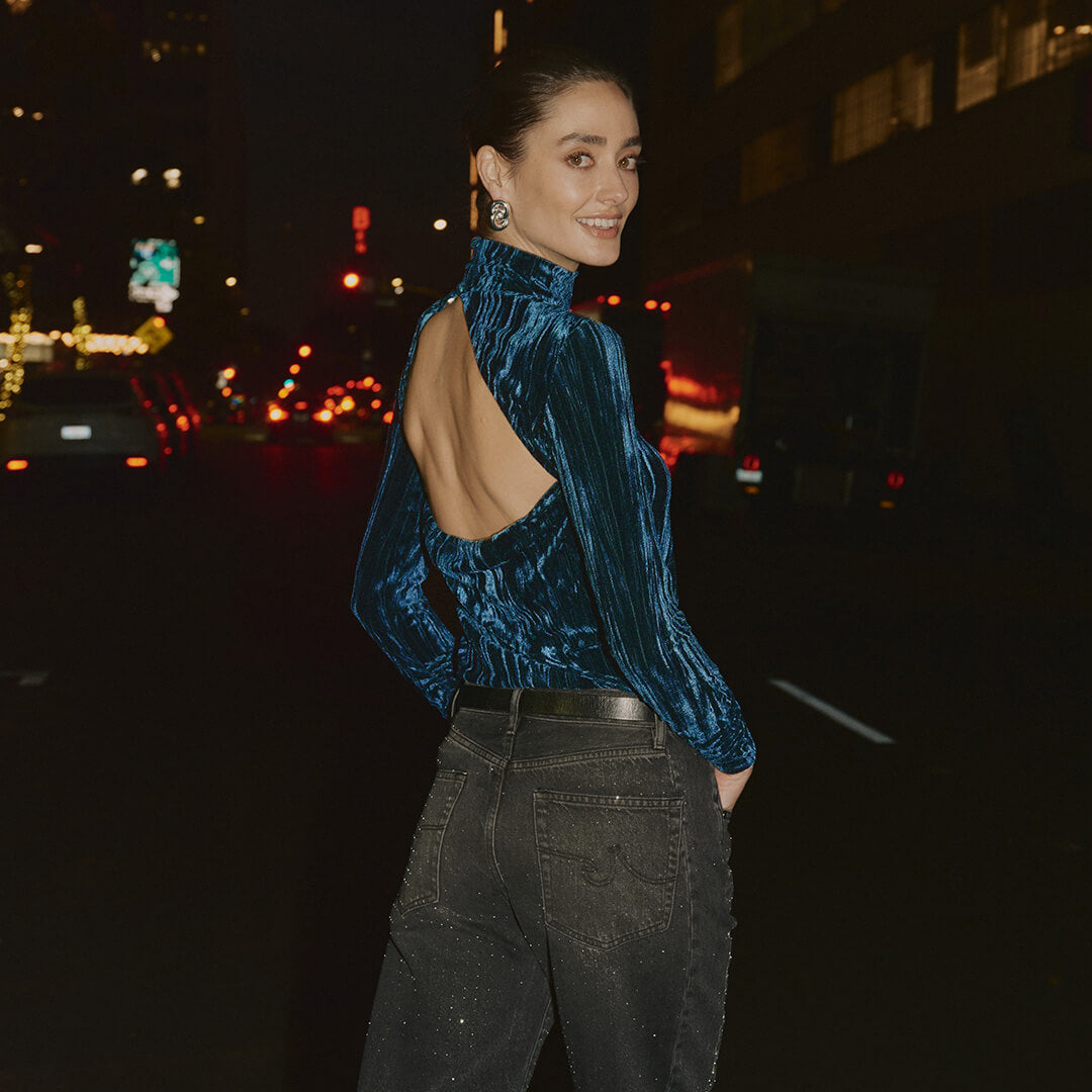 Woman wearing a blue velvet top with an open back and dark jeans on a city street at night.