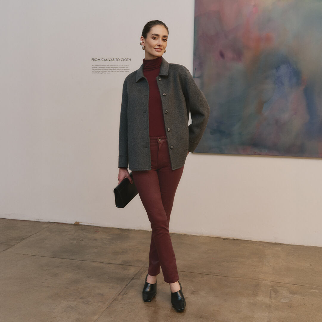Woman in a gray coat and red pants standing in an art gallery with a colorful painting in the background.