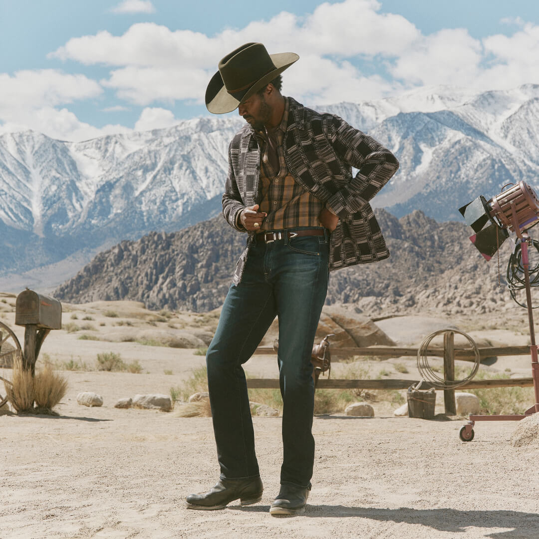Man in cowboy hat and plaid shirt standing in a desert with mountains in the background