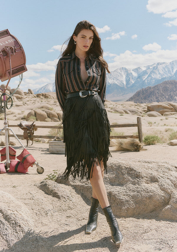 Woman in a stylish outfit with a fringe skirt and boots in a desert setting with mountains in the background.