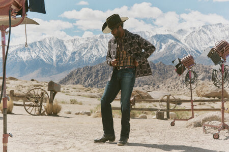 Man in cowboy hat and plaid shirt standing in a desert with mountains in the background