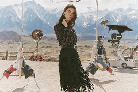 Woman in a black fringed skirt standing in front of a mountainous landscape with film equipment around.