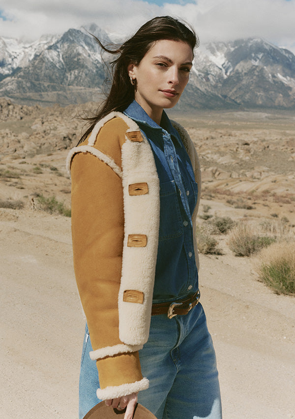 Woman wearing a brown shearling jacket in a desert landscape with mountains.