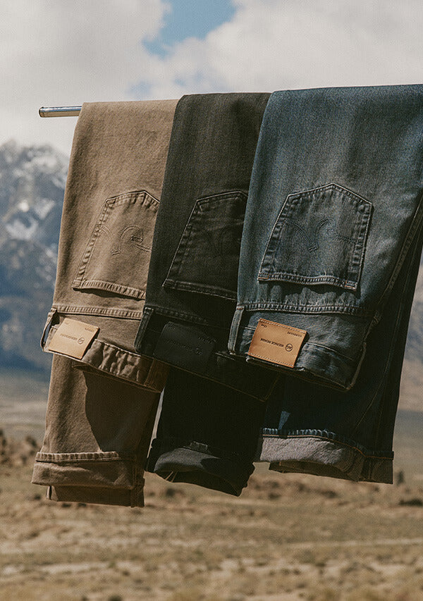 Three pairs of jeans in brown, black, and blue hanging on a clothesline with a mountainous background.