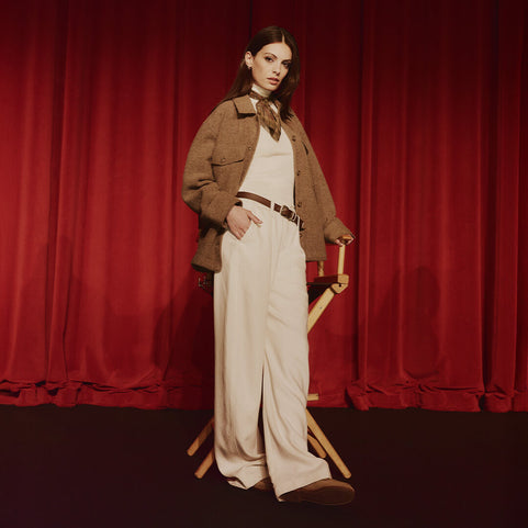 Woman in a brown jacket and white pants standing next to a director's chair with a red curtain background