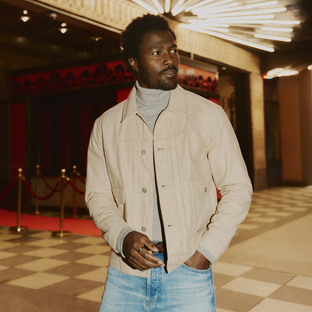 Man standing in a stylish interior setting wearing a beige jacket and blue jeans.