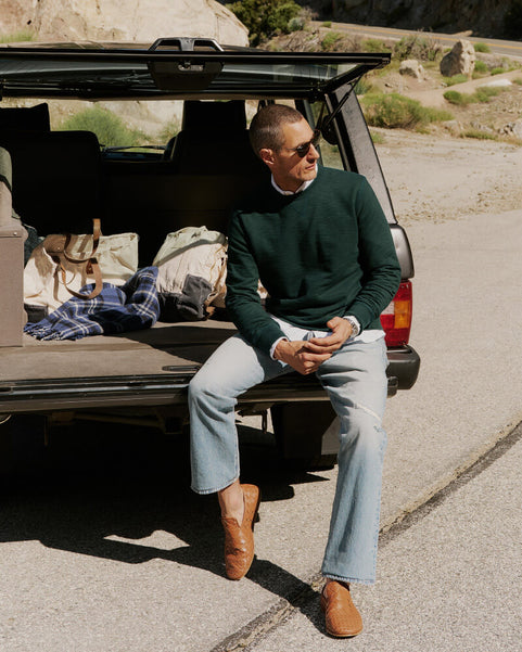 Man sitting on a car trunk with open doors, wearing sunglasses and a green sweater.