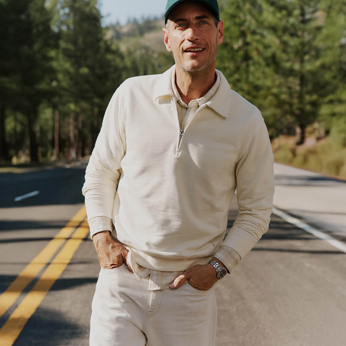 Man in a cream sweater and cap standing on a road with trees in the background