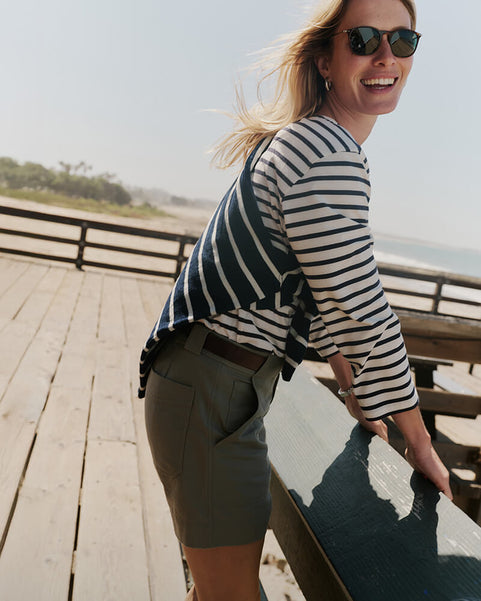 Woman in a striped shirt and shorts standing on a wooden boardwalk by the beach.