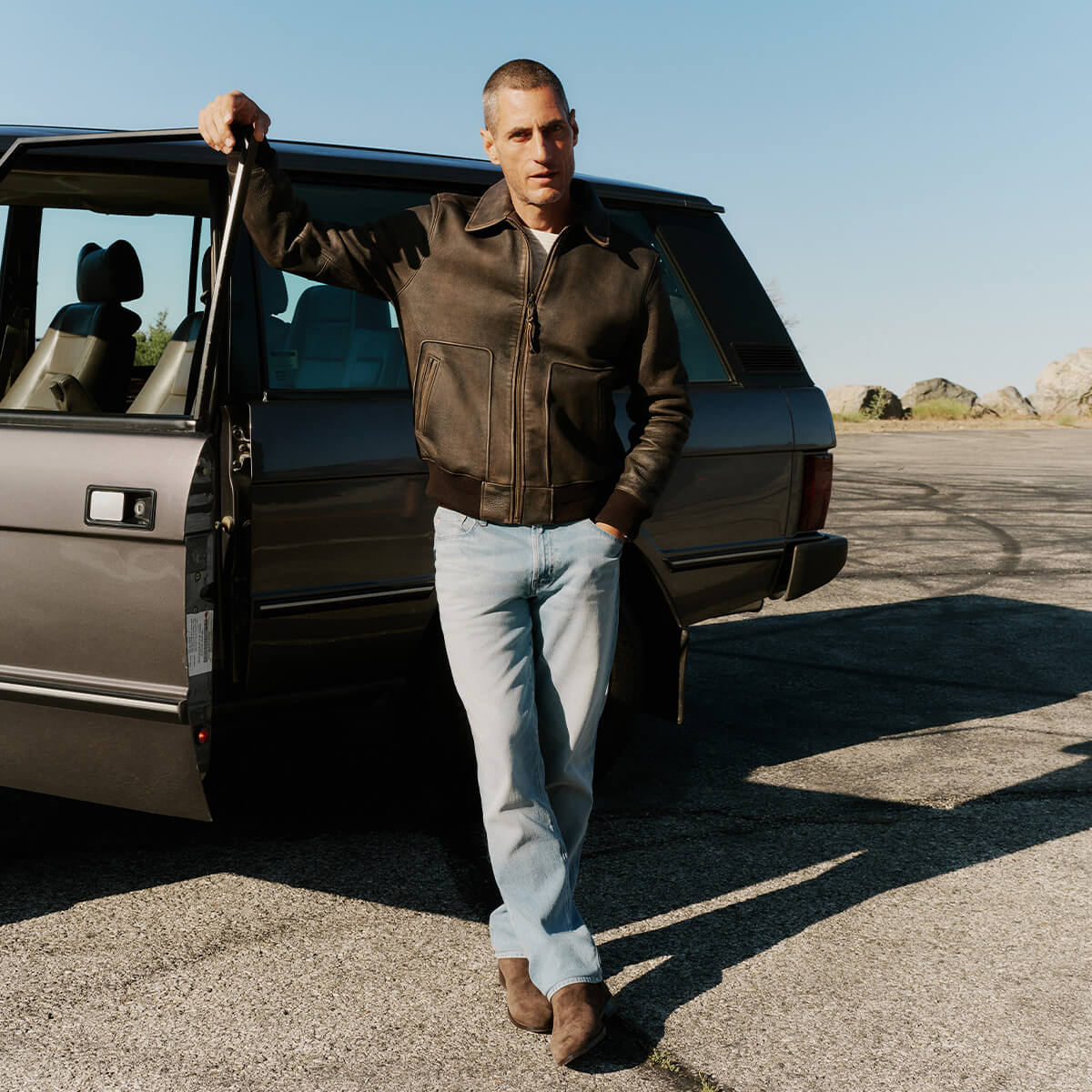 Man in a leather jacket standing next to an old-fashioned car in a parking lot.