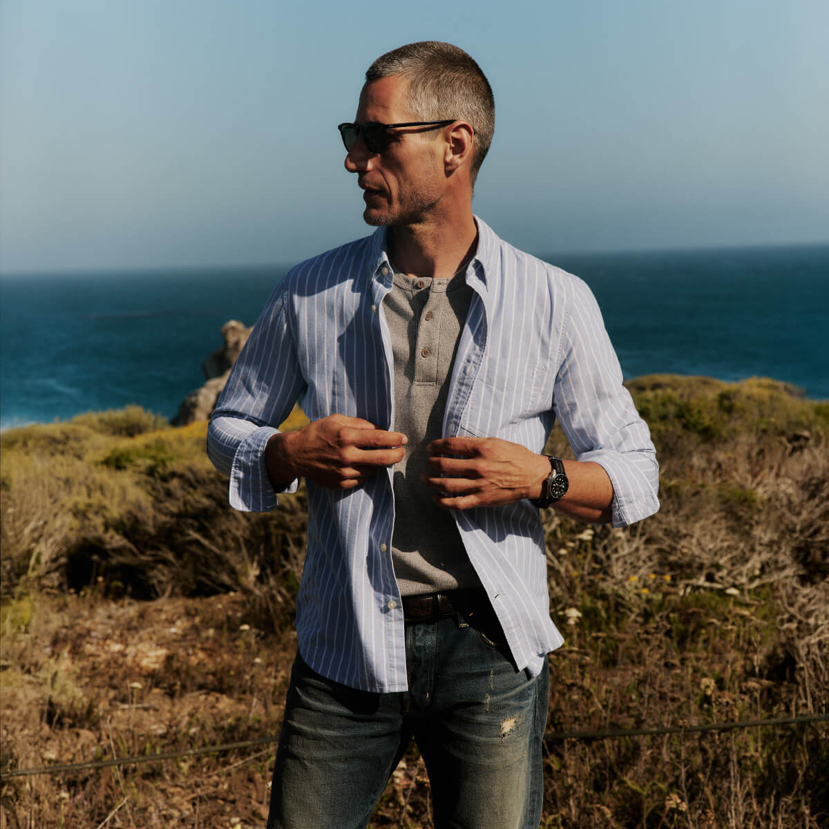 Man standing on a cliff overlooking the ocean wearing sunglasses and a striped shirt.