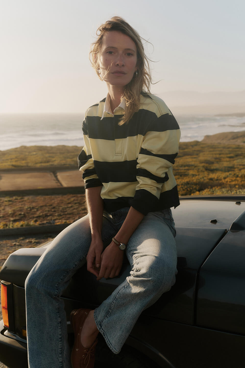Person sitting on a car by a scenic coastal area wearing a striped shirt and jeans.
