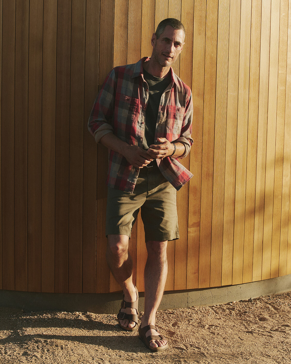 Man wearing a plaid shirt and shorts standing against a wooden wall.