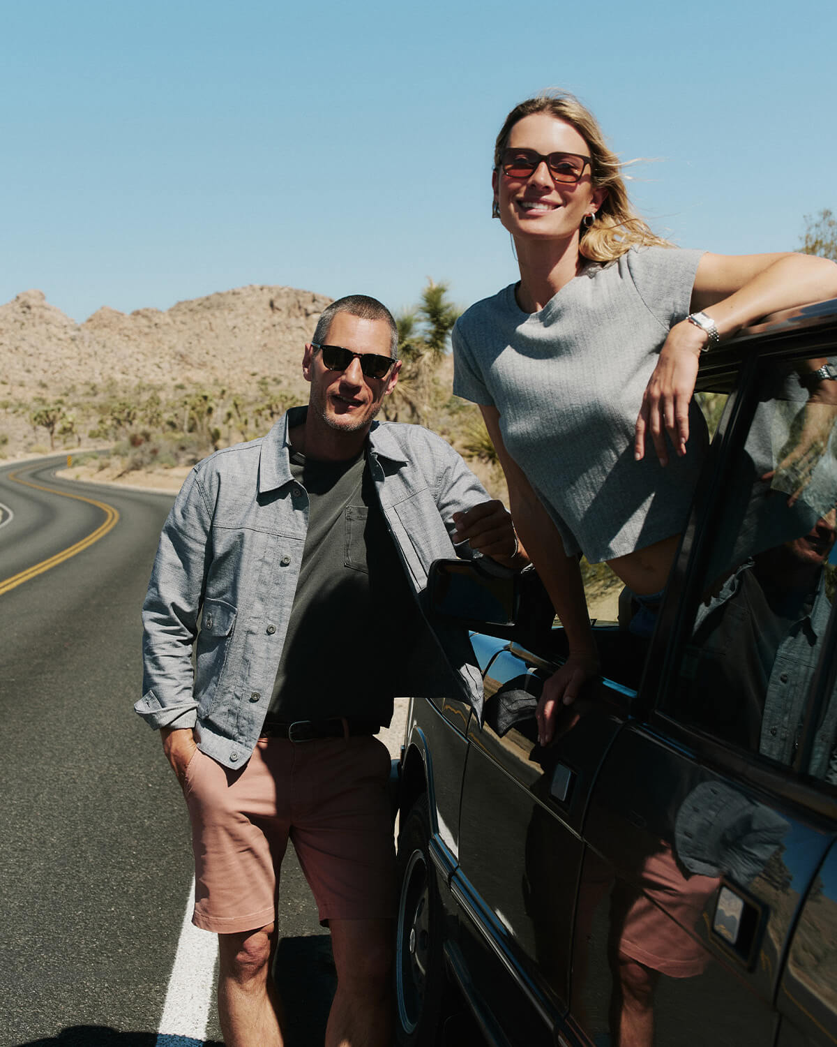 Two people standing by a car on a road with mountains in the background