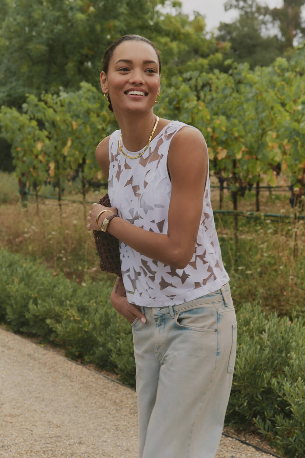 Woman standing in a vineyard wearing a white sleeveless top and light blue jeans.