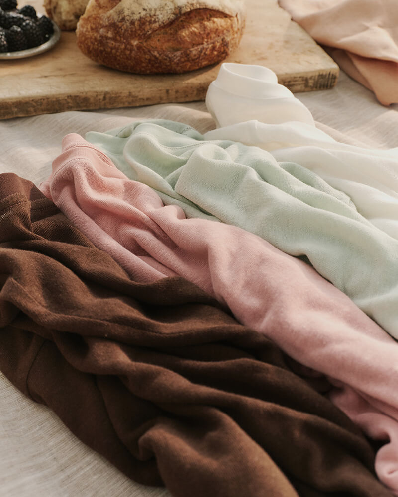 Stack of folded fabric in brown, pink, and green on a wooden surface with bread in the background.