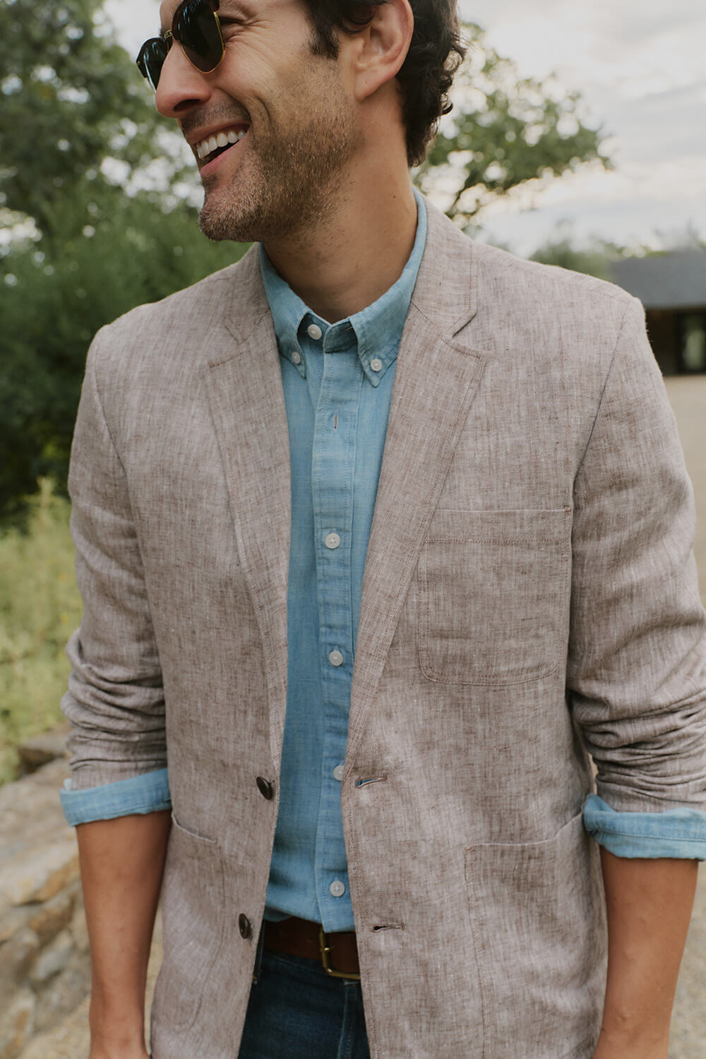 Man wearing a light brown blazer over a blue shirt with sunglasses, standing outdoors.