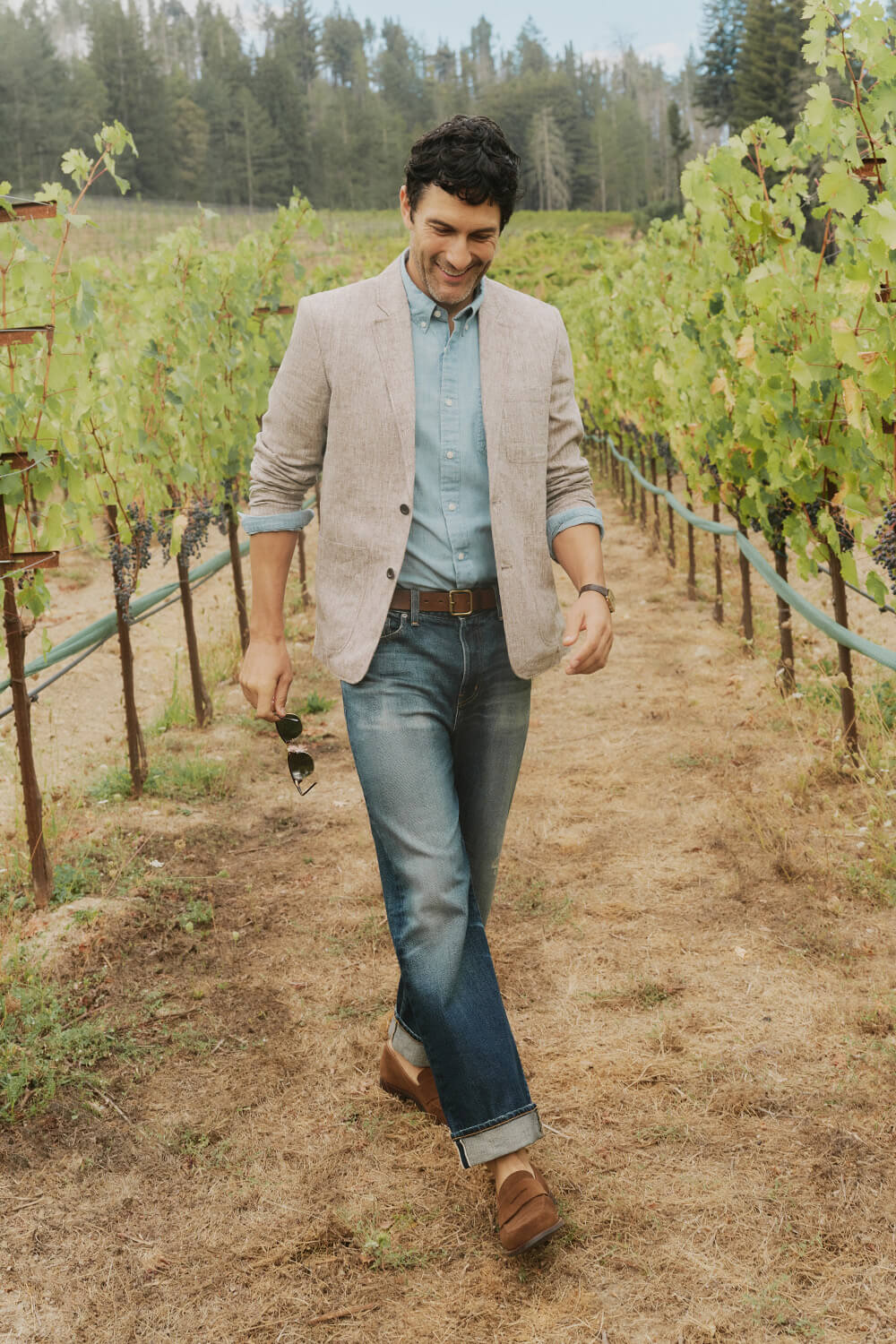 Man walking through a vineyard wearing a beige blazer and blue jeans.