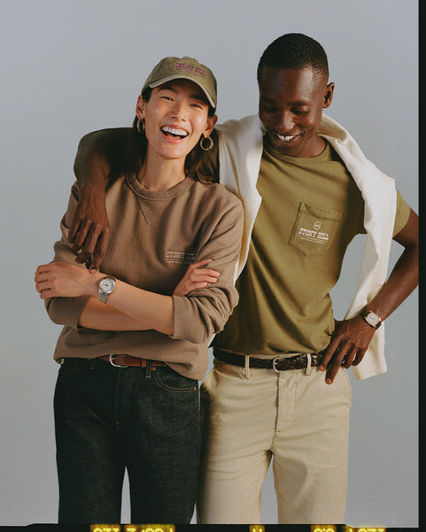 Two people standing together, one wearing a brown sweatshirt and cap, the other in a green shirt and beige pants, on a gray background.
