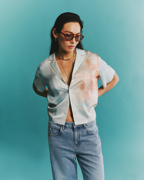 Woman wearing a light-colored blouse with a floral pattern over a white top, paired with high-waisted jeans against a teal background.