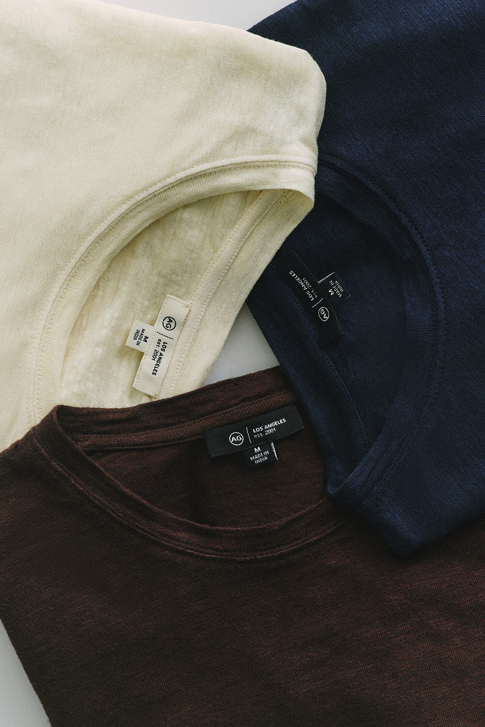 Three folded t-shirts in beige, brown, and navy on a white surface.