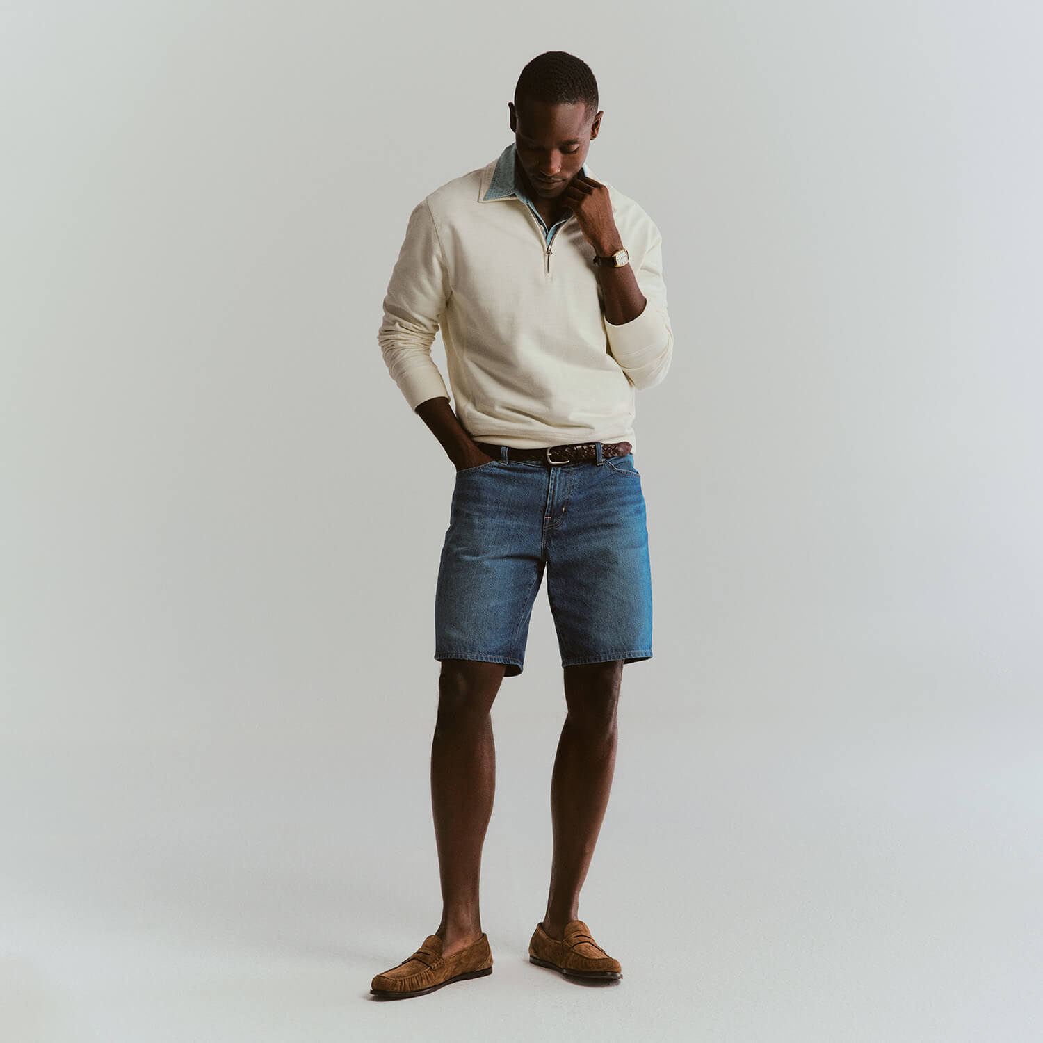 Man wearing a cream sweater, blue denim shorts, and brown shoes on a plain background