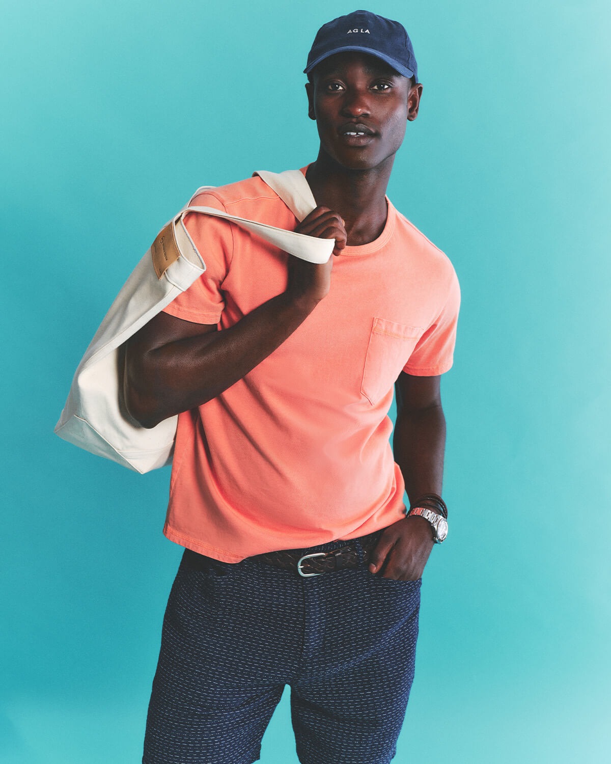 Man wearing a coral shirt and navy cap holding a white bag against a teal background