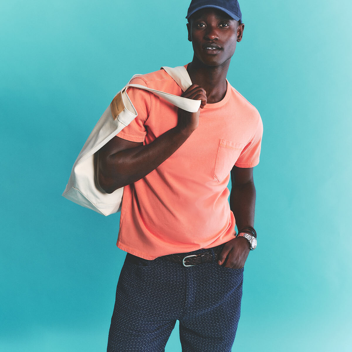 Man wearing a pink shirt and blue cap holding a white bag against a teal background
