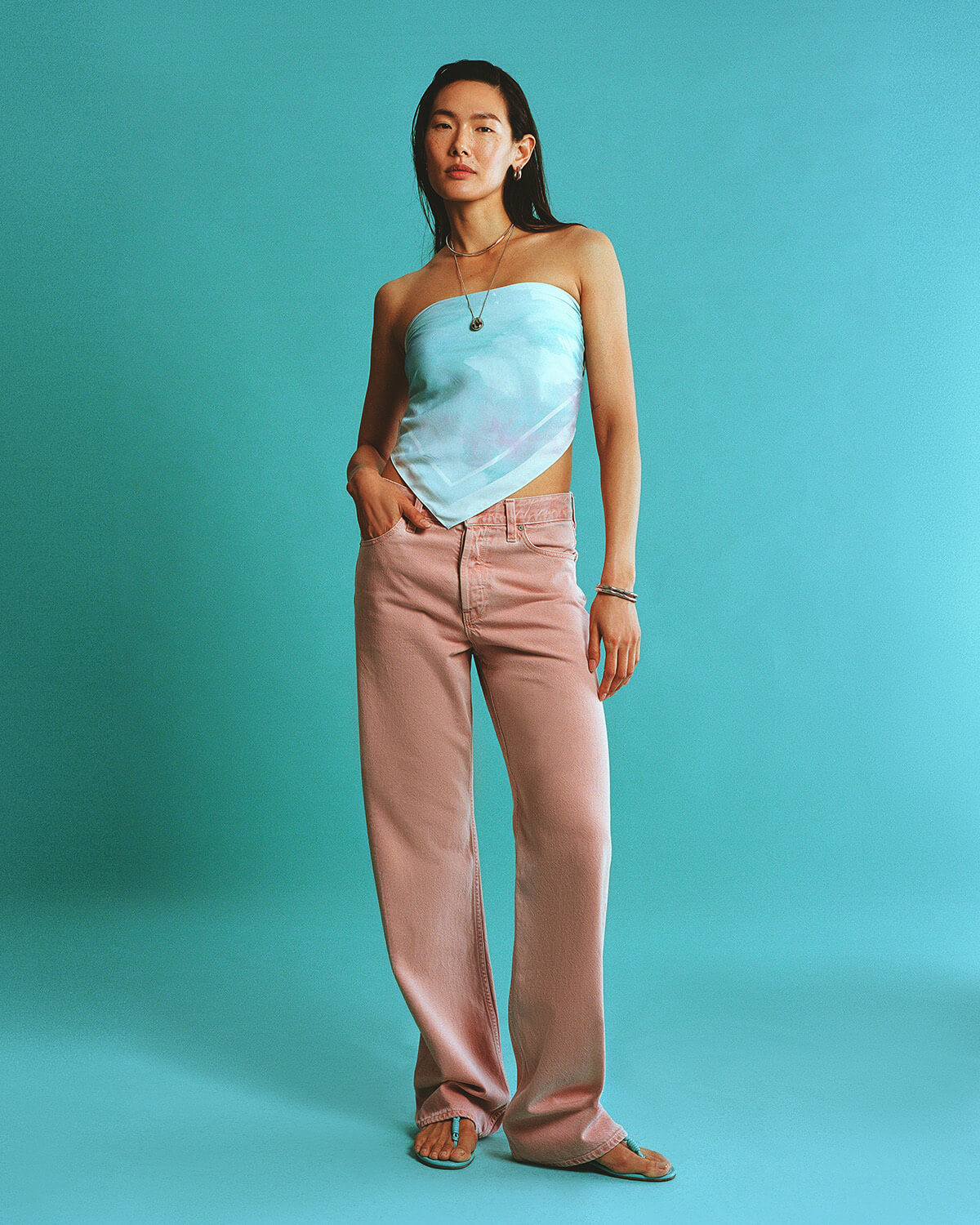 Woman wearing a light blue strapless top and pink pants against a teal background