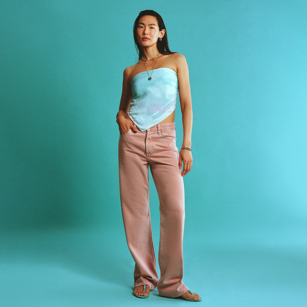 Woman wearing a light blue strapless top and pink pants against a teal background