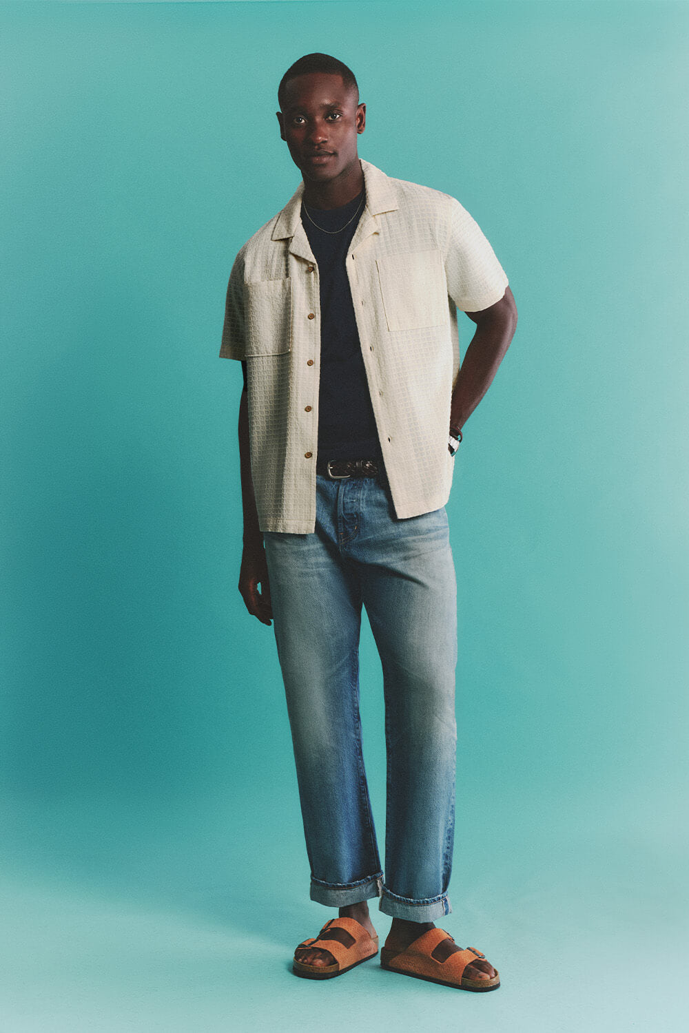 Man wearing a white shirt, black t-shirt, blue jeans, and brown sandals against a teal background
