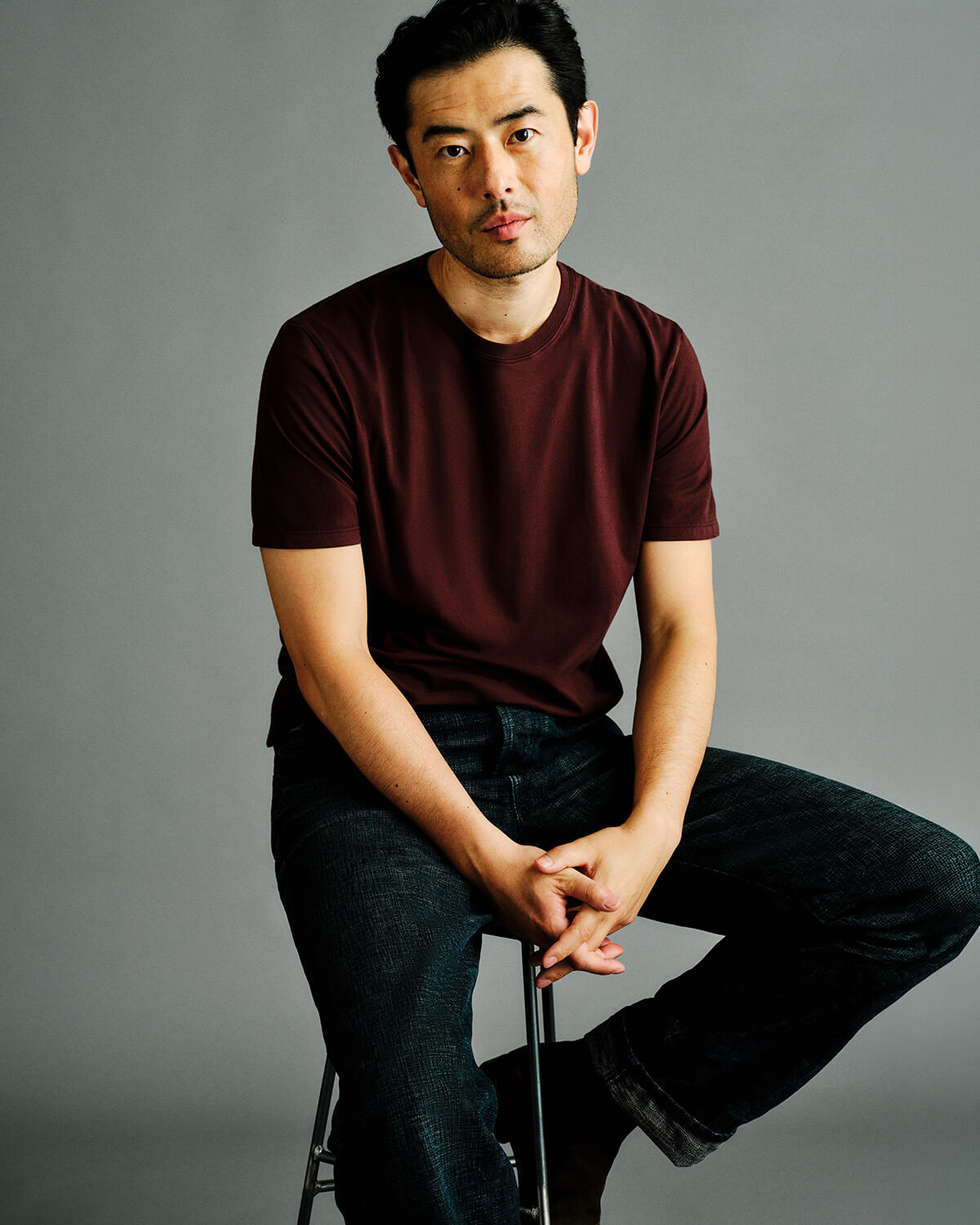 A man wearing a dark maroon crewneck t-shirt and dark jeans, sitting on a stool against a solid gray studio background.