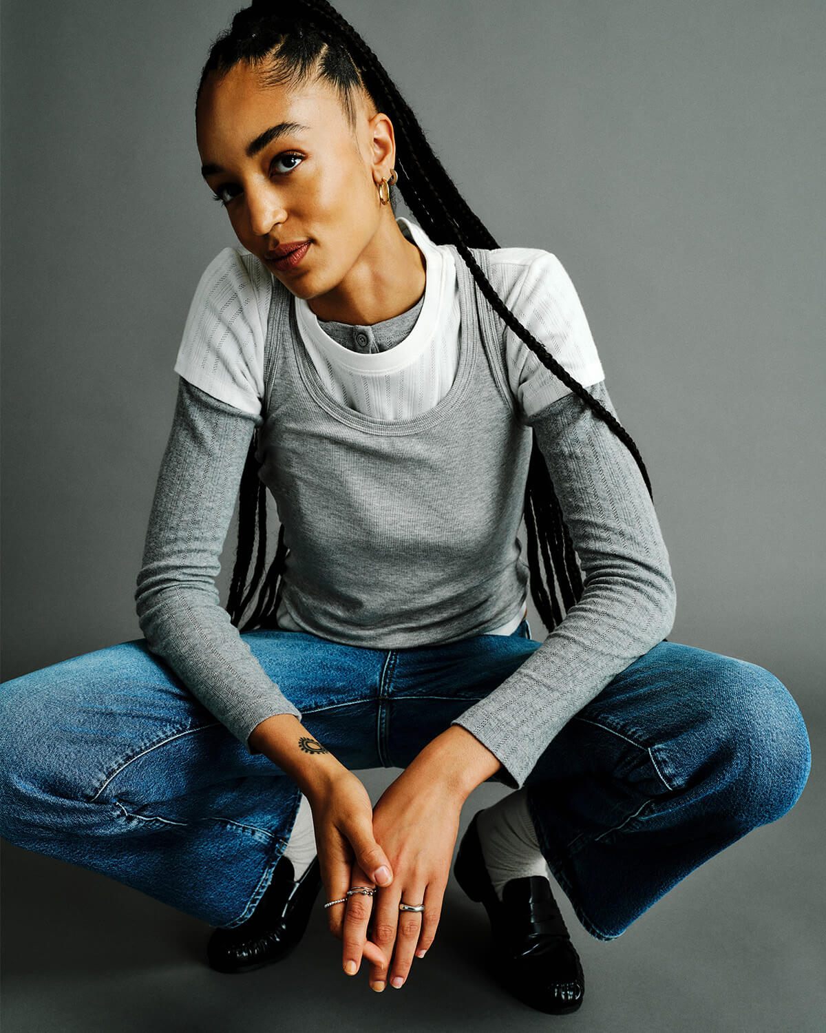 A model crouching in a studio setting, wearing a layered gray and white long-sleeve top, blue jeans, white socks, and black loafers against a solid gray background.