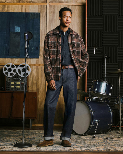 A man stands in a music studio wearing a brown and black plaid flannel shacket, a dark denim shirt, and selvage jeans. A vintage microphone, reel-to-reel machine, and drum set are visible in the room.