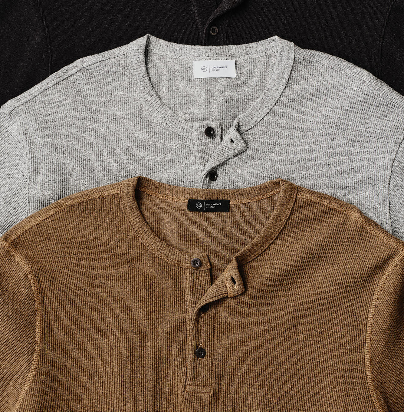 Three ribbed henley shirts layered in a flat-lay: a tan/brown shirt in the foreground and a light gray shirt behind it, with a dark shirt partially visible at the top edge.