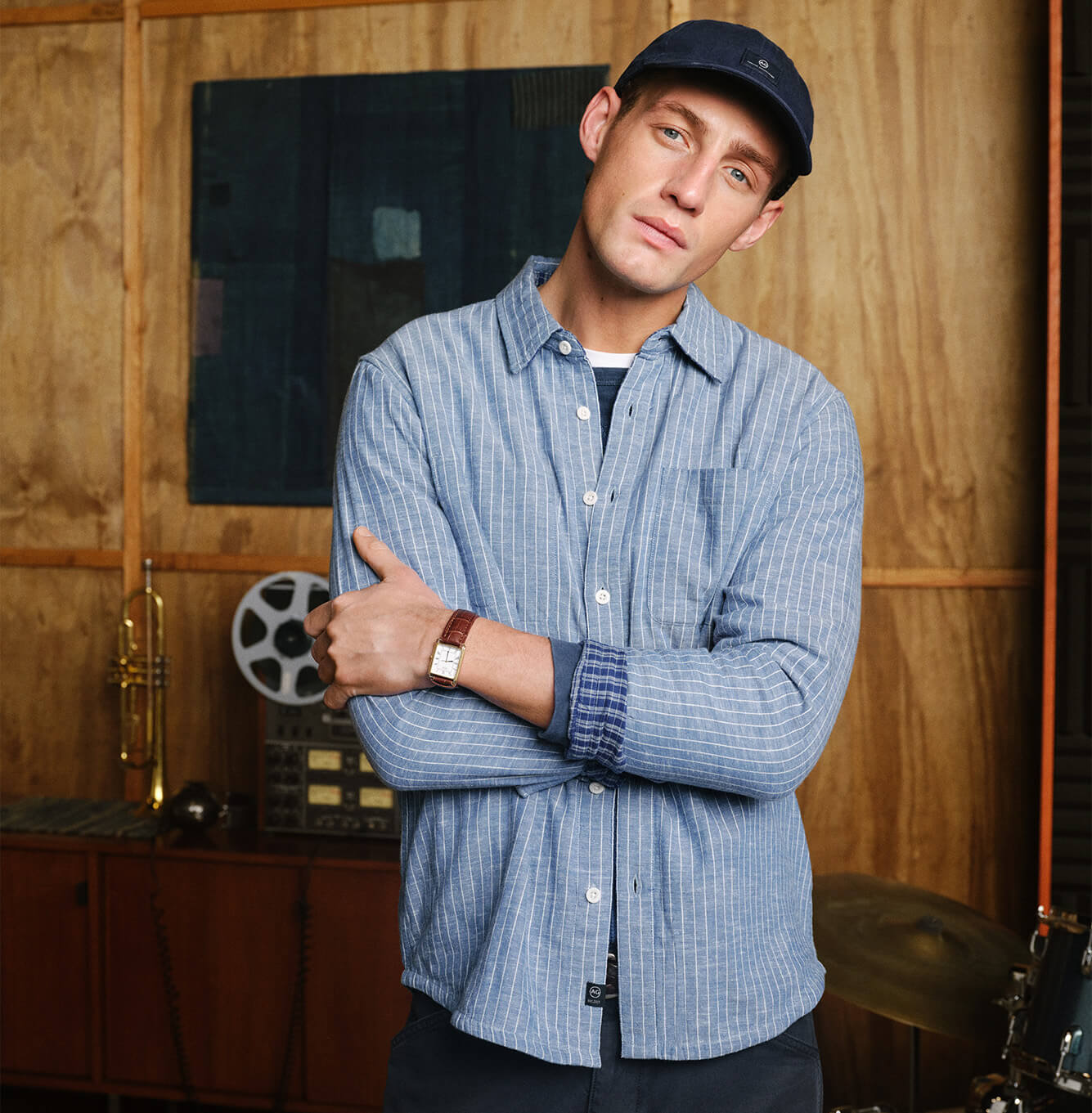 A man wearing a blue and white vertically striped button-up shirt, a navy AG cap, standing with arms crossed in front of a wood-paneled wall with vintage audio equipment.