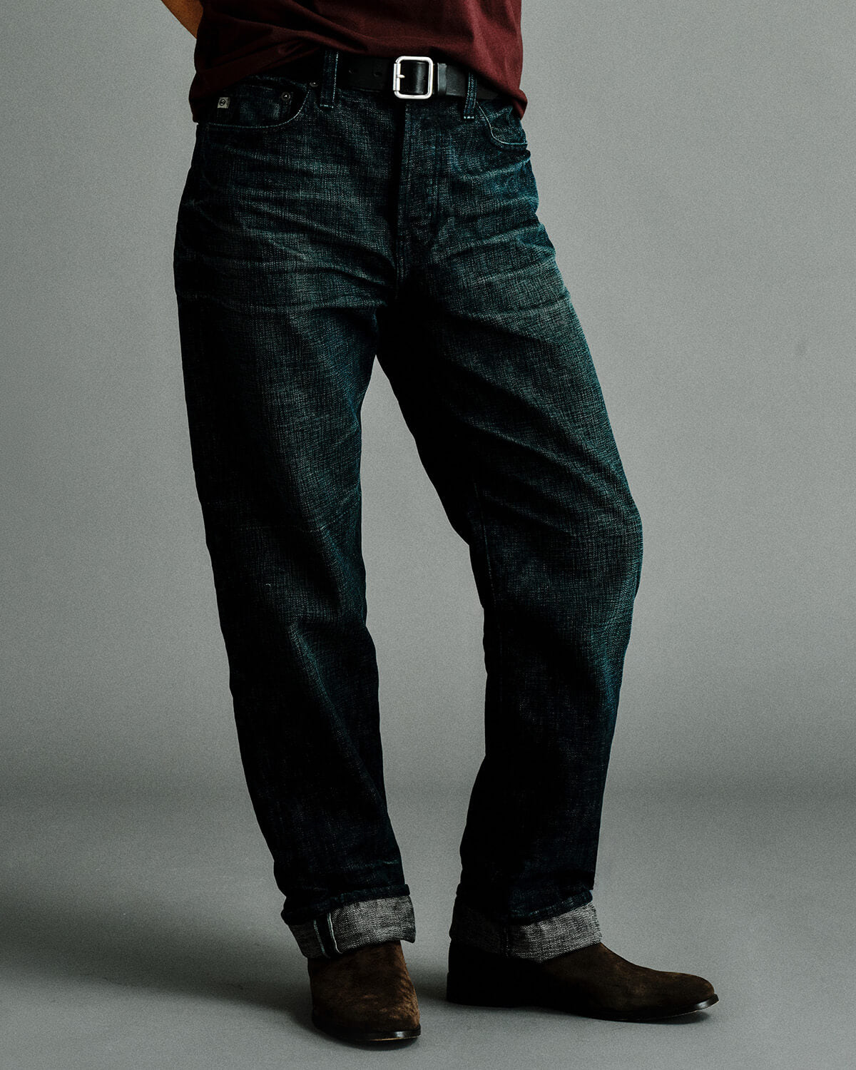 A person wearing dark indigo denim jeans with a silver buckle belt, a burgundy shirt, and brown boots, shown from the waist down against a plain gray studio background.