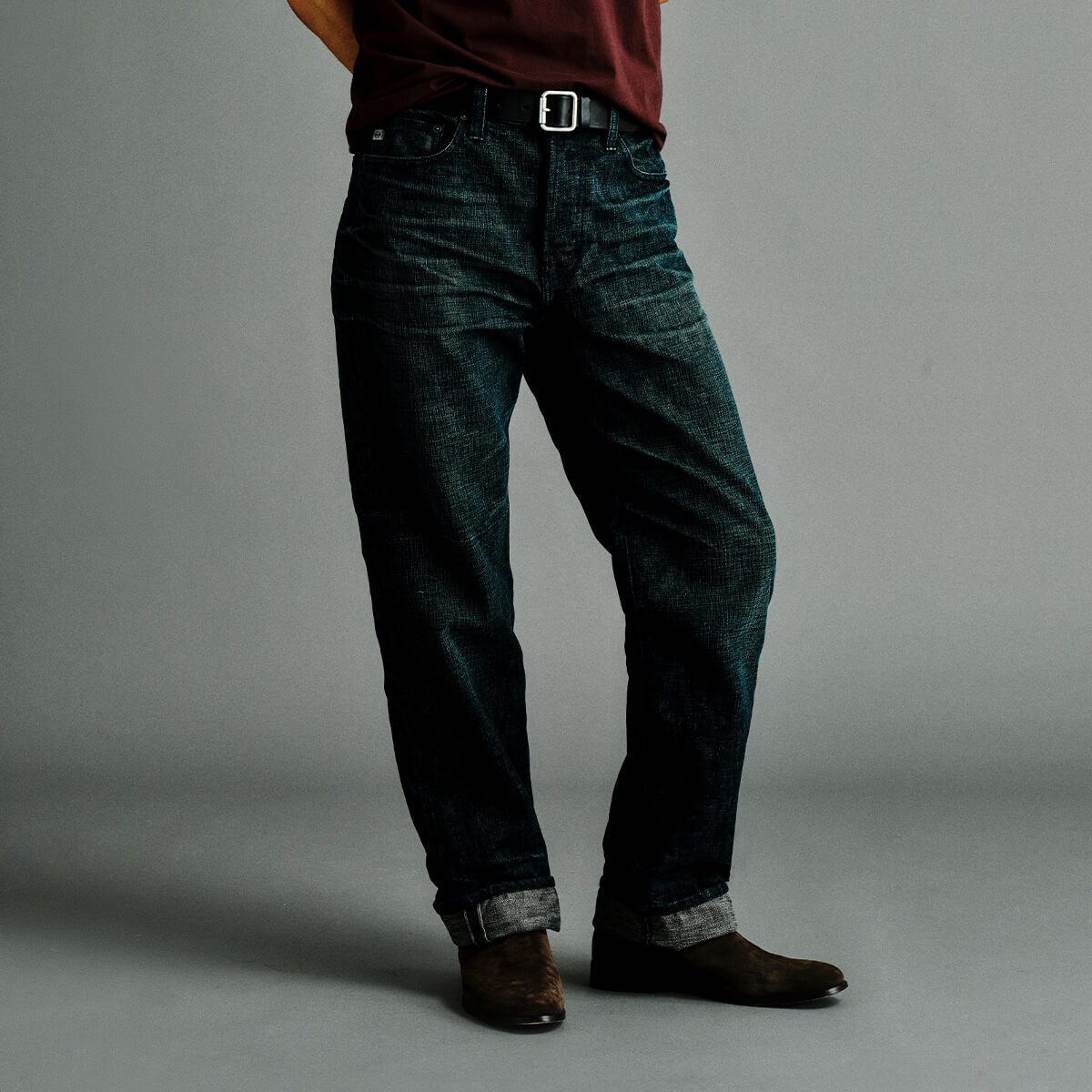 A person wearing dark indigo denim jeans with rolled cuffs, paired with brown boots and a dark red shirt, standing against a plain gray studio background.